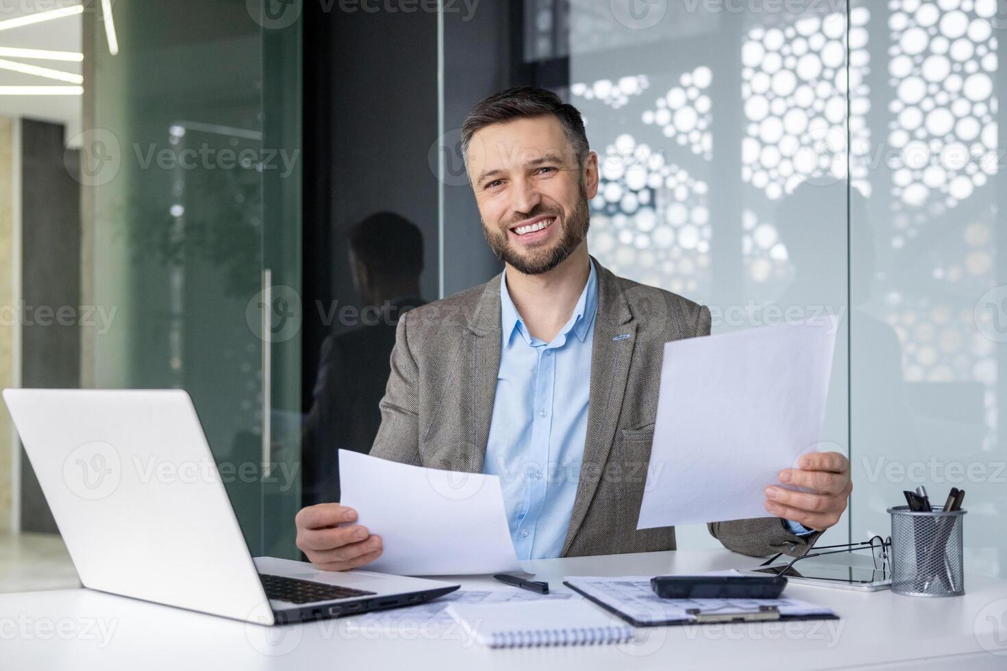 Confident businessman sitting at a desk with a laptop, holding documents, and smiling in a modern glass office. Concept of successful work and professional achievement. photo
