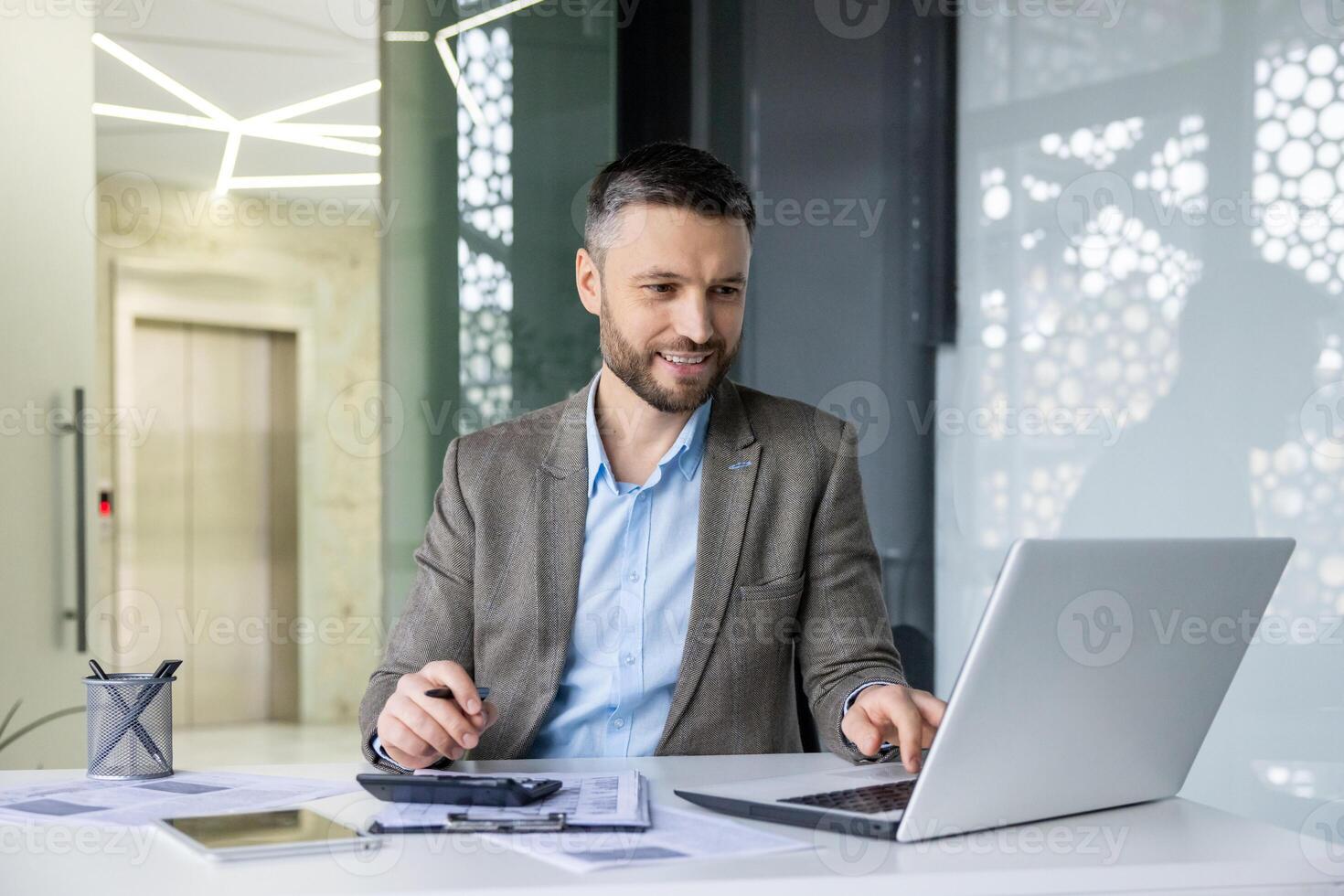 Smiling businessman working on laptop in modern office, analyzing documents and taking notes. Professional and focused in corporate environment. photo