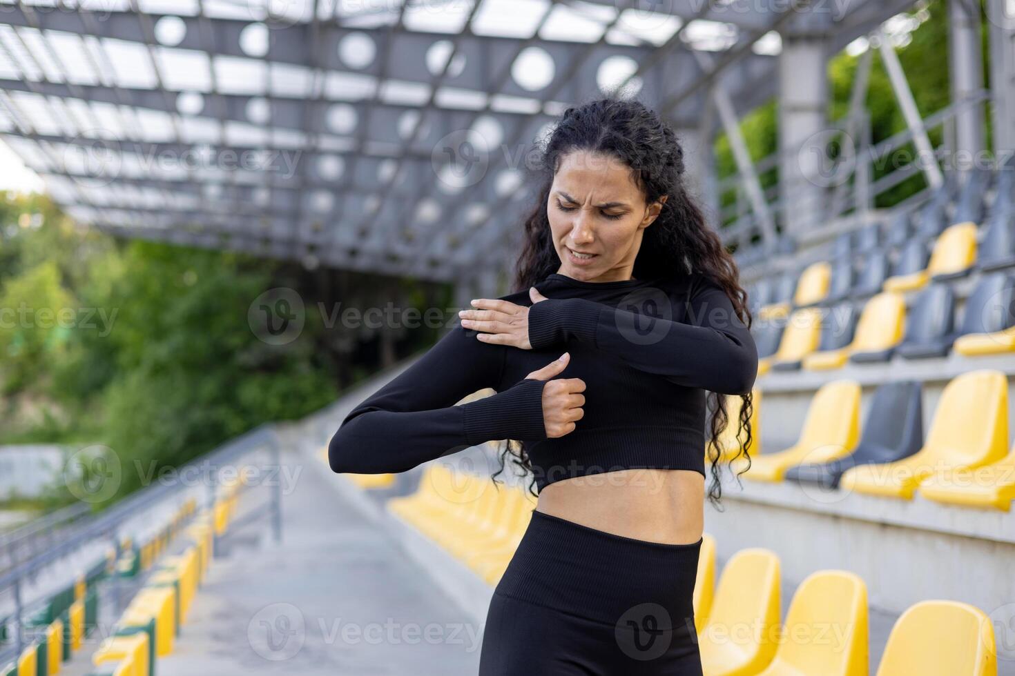 A hispanic woman experiences sharp pain in her shoulder while exercising at a stadium. Her expression shows discomfort and pain, highlighting a common sports injury. photo