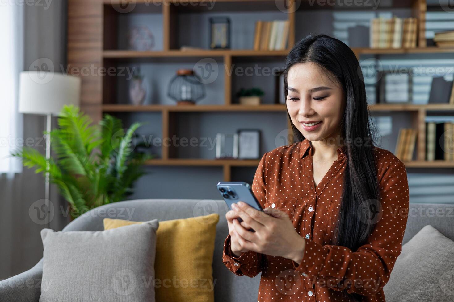 Asian woman smiling and using smartphone at home. Seated on sofa in modern living room, engaged in social media browsing or chatting. Captures moments of relaxation and connectivity. photo