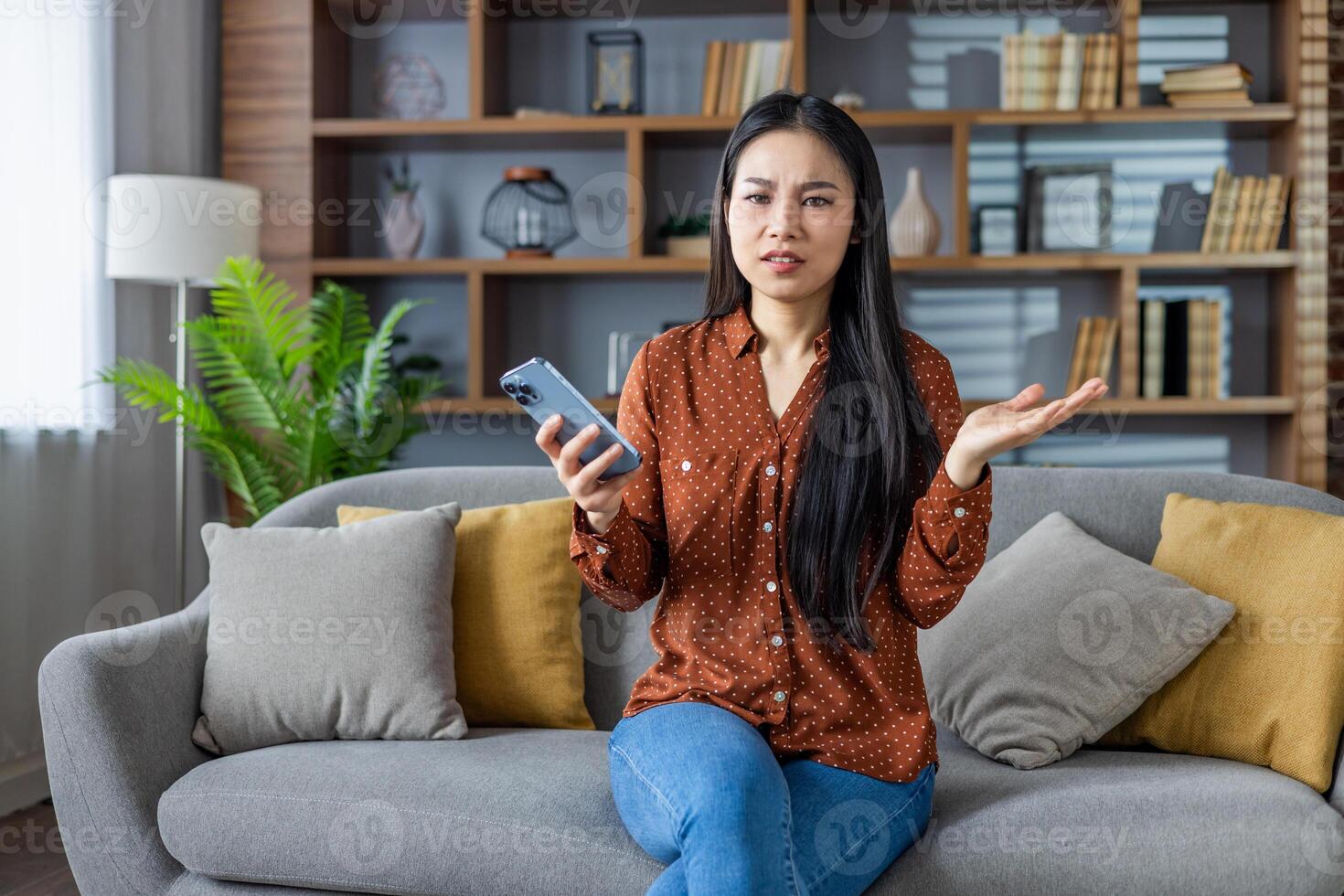Asian woman seated on sofa holding smartphone looks confused and frustrated, expressing bewilderment and doubt. Comfortable home environment with bookshelves in background adds a cozy touch. photo
