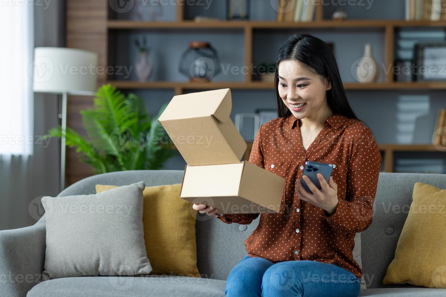 Asian woman sitting on sofa, happily opening delivered package with smartphone in hand. Expresses excitement from online shopping and receiving order at home. Represents joyful unboxing experience. photo