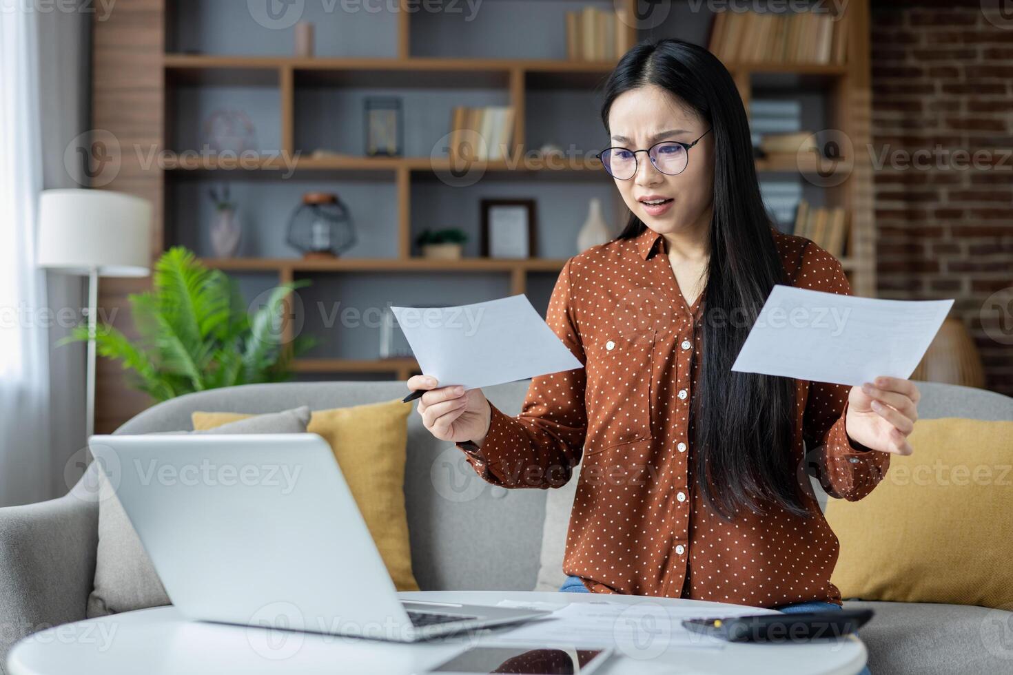 Asian woman analyzing papers at home, wearing glasses, using laptop. Focusing on important details, multitasking in living room. Concept of concentration, home office, paperwork management. photo