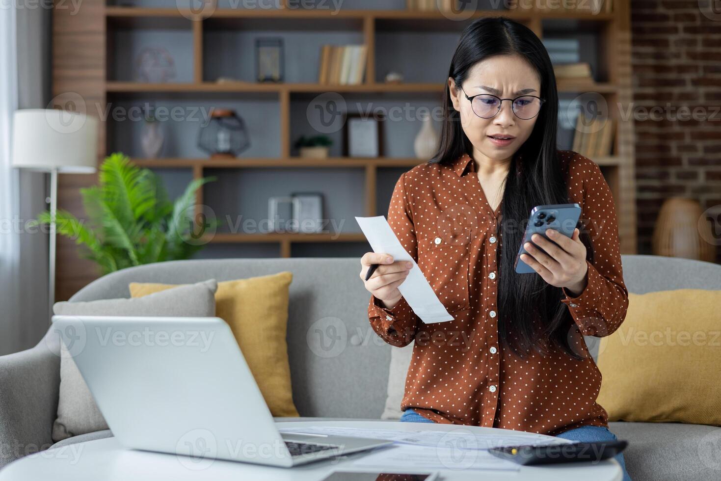 Asian woman feeling stressed while managing bills at home using smartphone and laptop. She holds bill with worried expression, immersed in online banking task. Concept of finance and technology. photo
