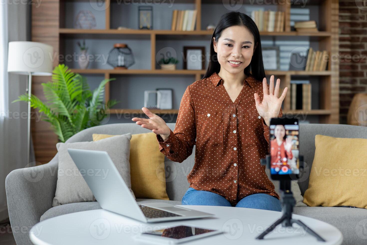 Asian woman engaging in conference at home. Sitting on sofa with laptop and phone tripod. Modern living room setting. Concept of remote work, online communication. photo