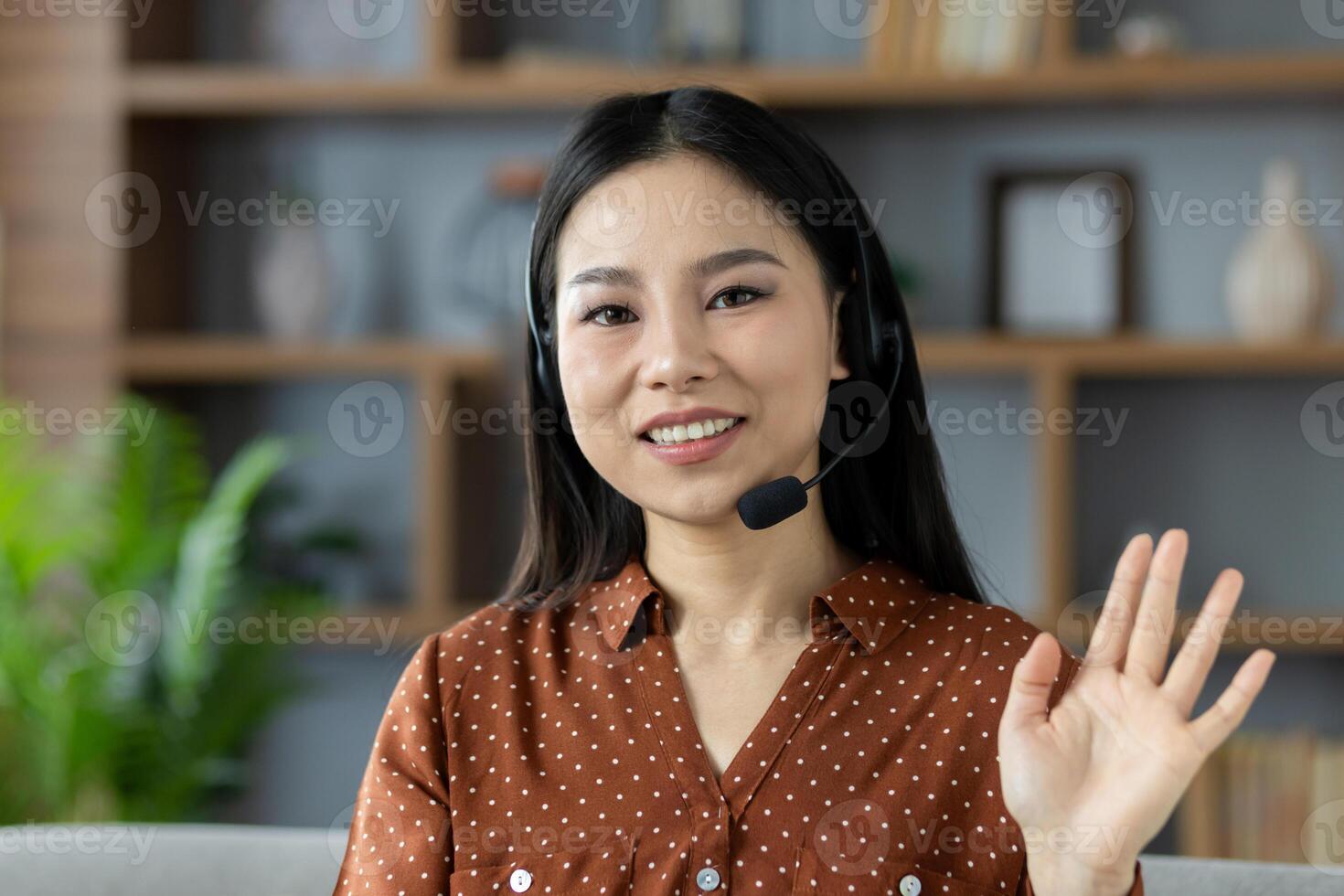 Cheerful Asian woman wearing headset, participating in online communication from comfortable setting, waves with friendly gesture. Image captures remote work, technology, connection, comfort, interaction. photo