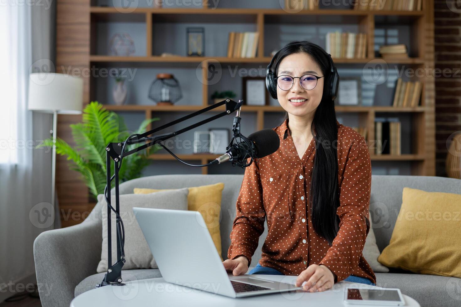 Asian woman hosting podcast in home studio, seated on sofa with laptop and microphone. Headphones on, conveying technology and communication in cozy living space with home office vibes. photo