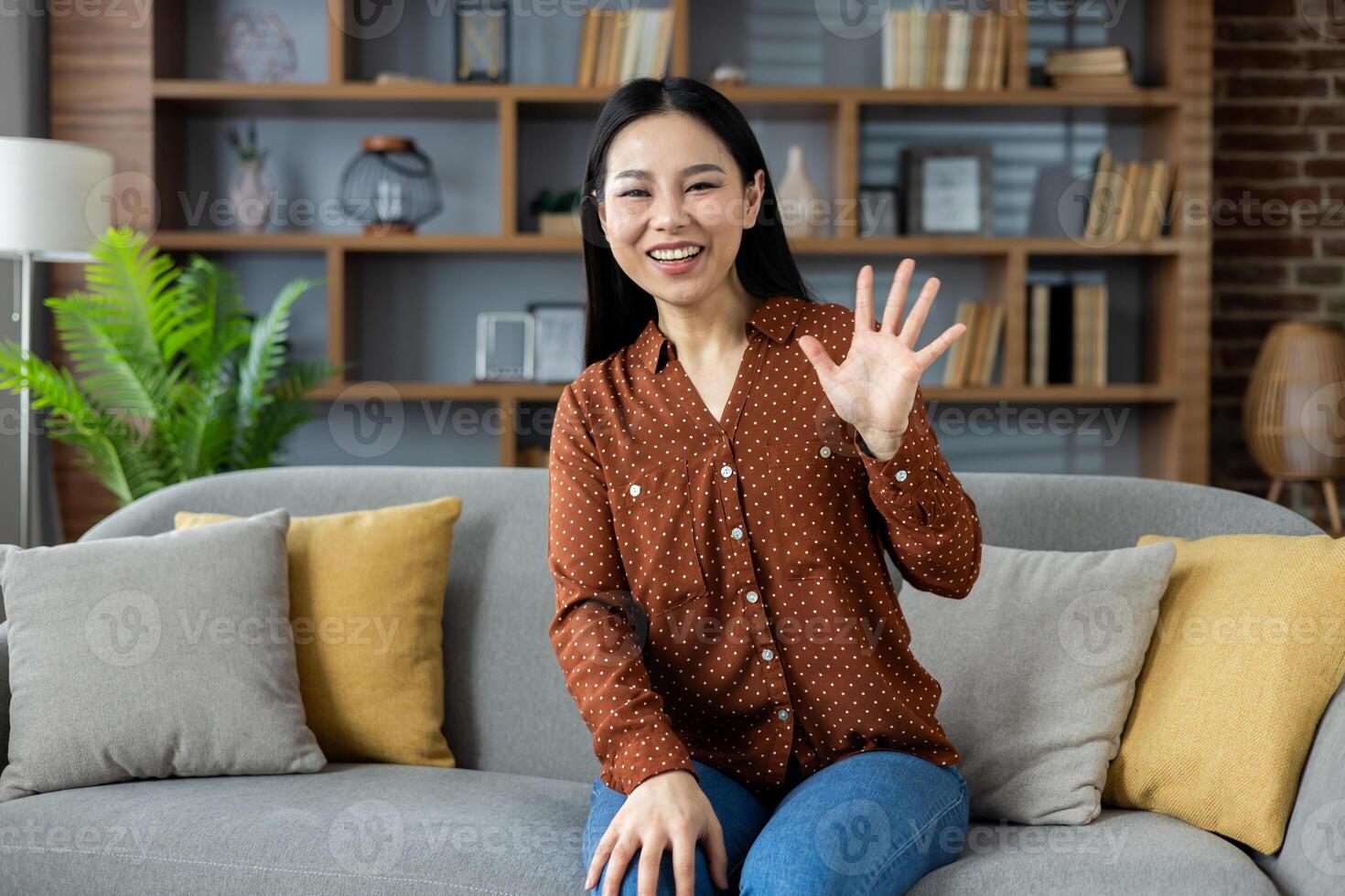 Asian woman smiling and waving while seated on sofa in modern home. She is dressed casually and appears happy. Room features include stylish shelves and comfortable furnishings. photo