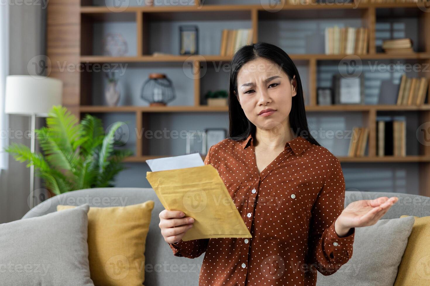 Asian woman with perplexed look holds envelope, expressing confusion over its contents. Scene captures emotion and curiosity in a cozy home environment, highlighting surprise. photo