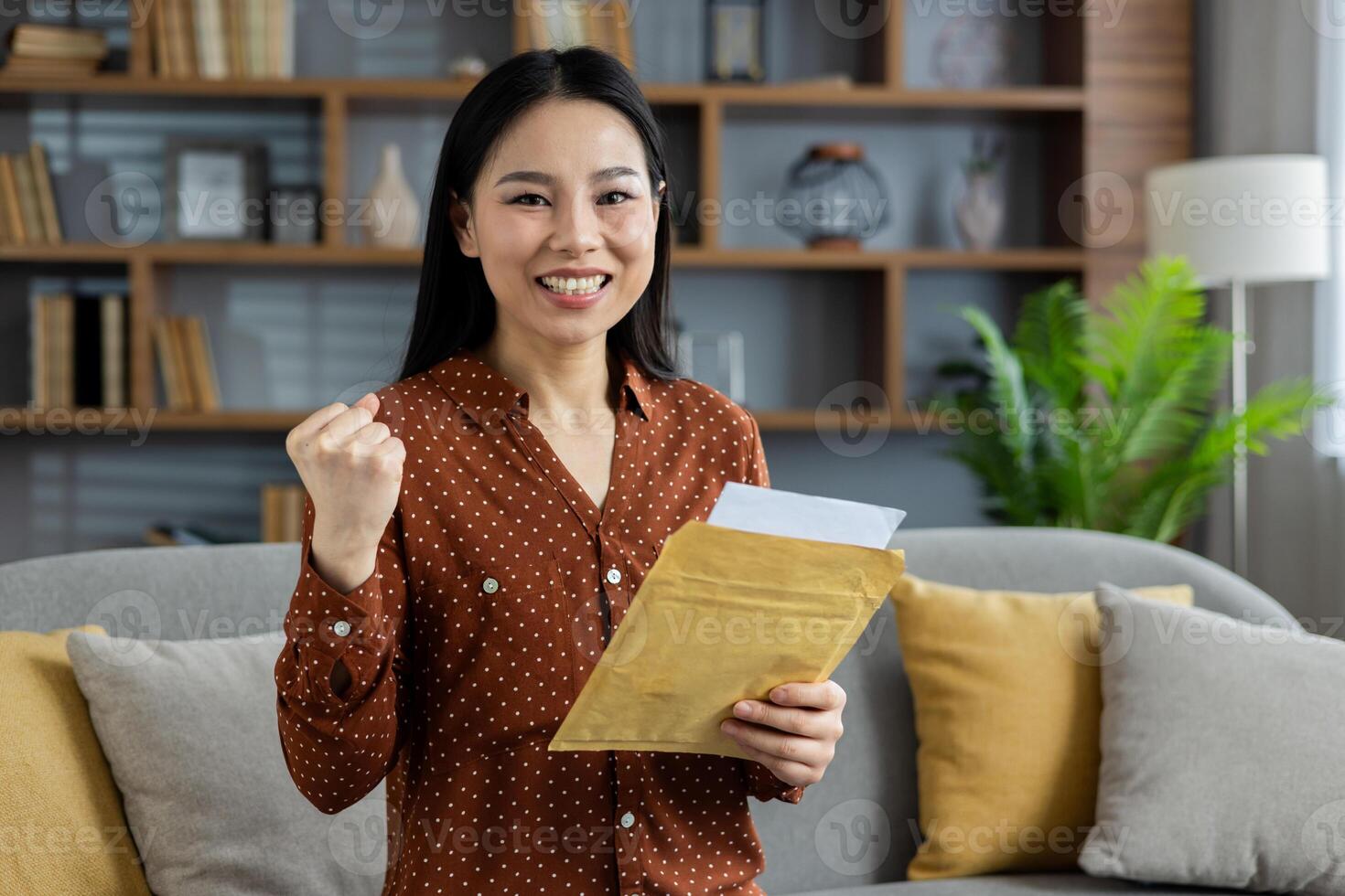 Asian woman expressing joy at home holding envelope in hand. Her happy and victorious expression represents success and positive news. Setting is cozy and comfortable. photo