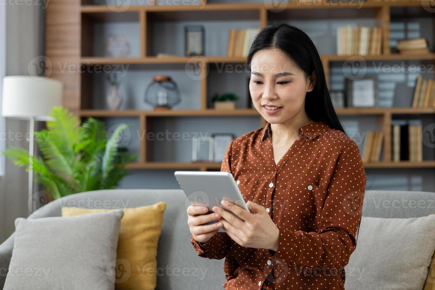 Asian woman using tablet at home. She is sitting on sofa, smiling, enjoying leisure time with technology. Casual wear enhances cozy atmosphere. Tablet and modern living space prominent. photo