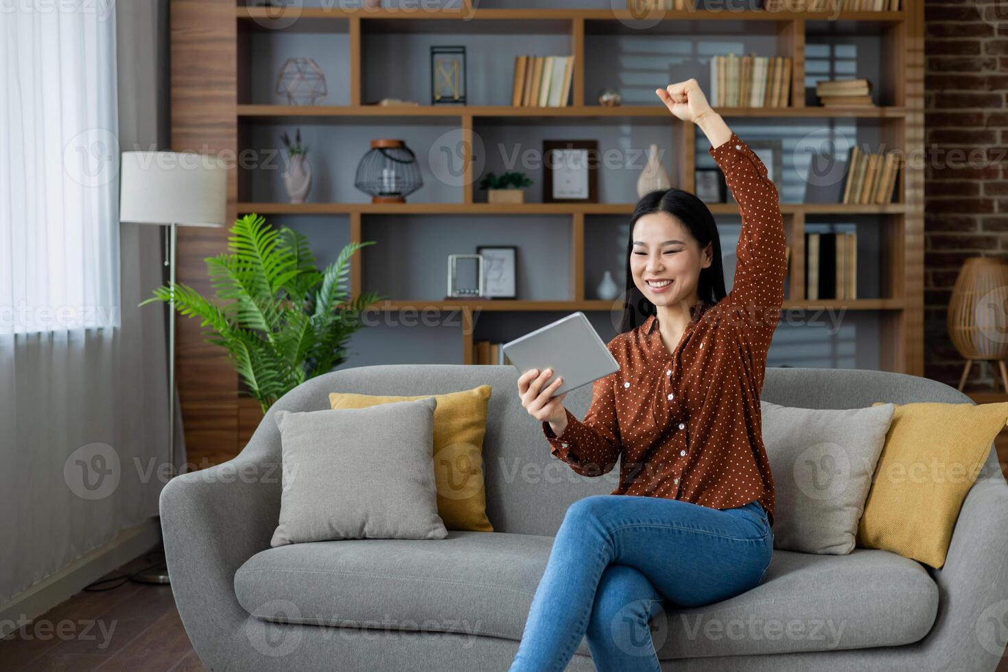 Asian woman sits on sofa using tablet, raising arm in celebration, expressing joy, success, and achievement. Ideal for concepts like happiness, technology, and work from home lifestyle. photo