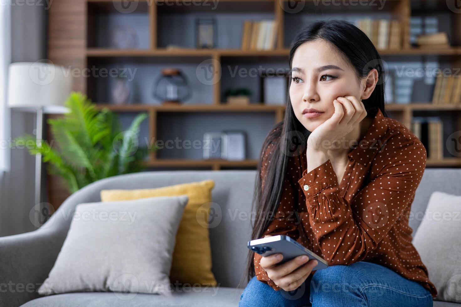 Young Asian woman sits on couch, holding phone, looking thoughtful. Casual attire suggests relaxed setting. Expression indicates contemplation or sadness, perhaps personal reflection at home. photo