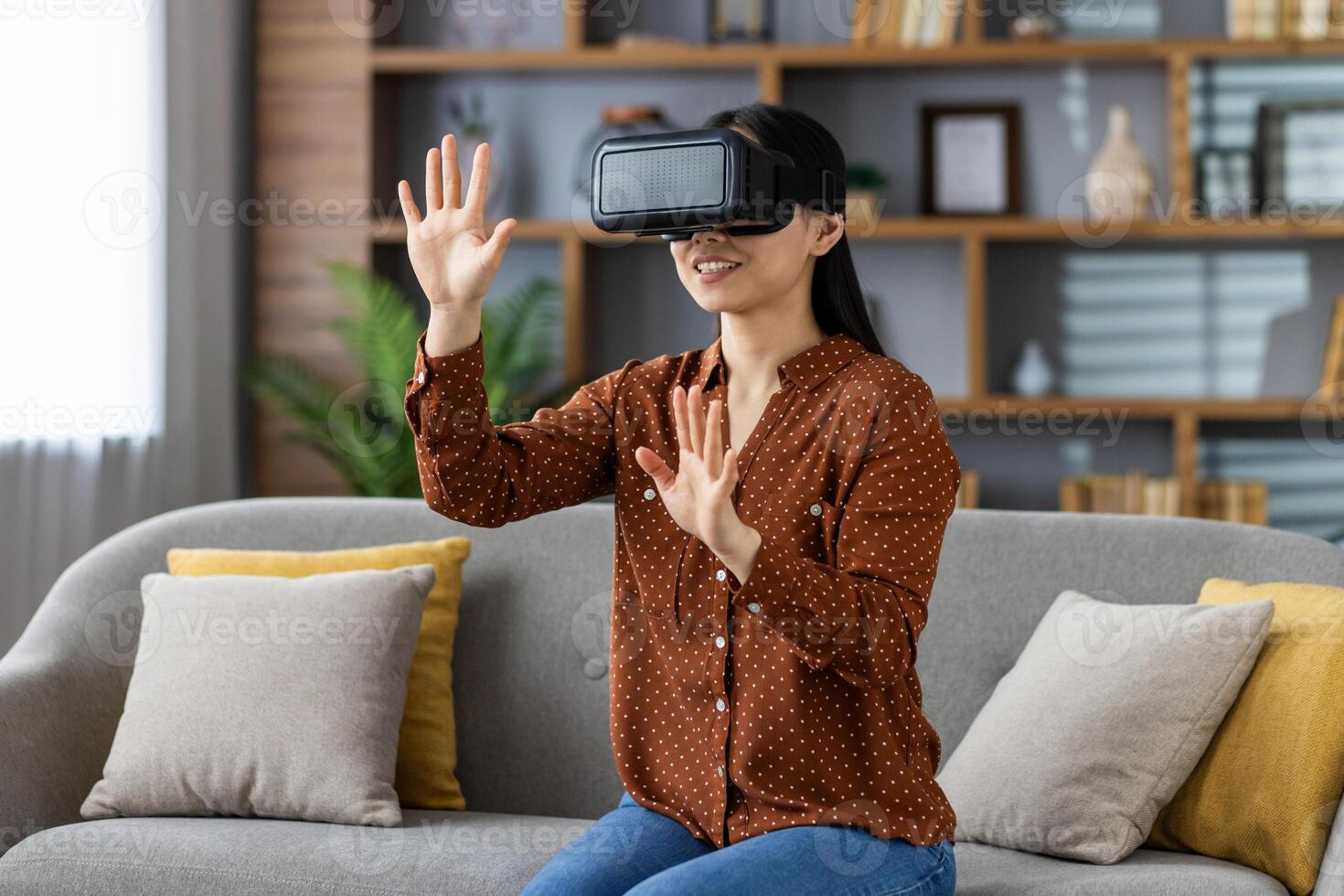 Asian woman explores virtual reality with VR headset at home. Seated on sofa, she's interacting with immersive digital content. Experience portrays joy, technology, and home comfort. photo