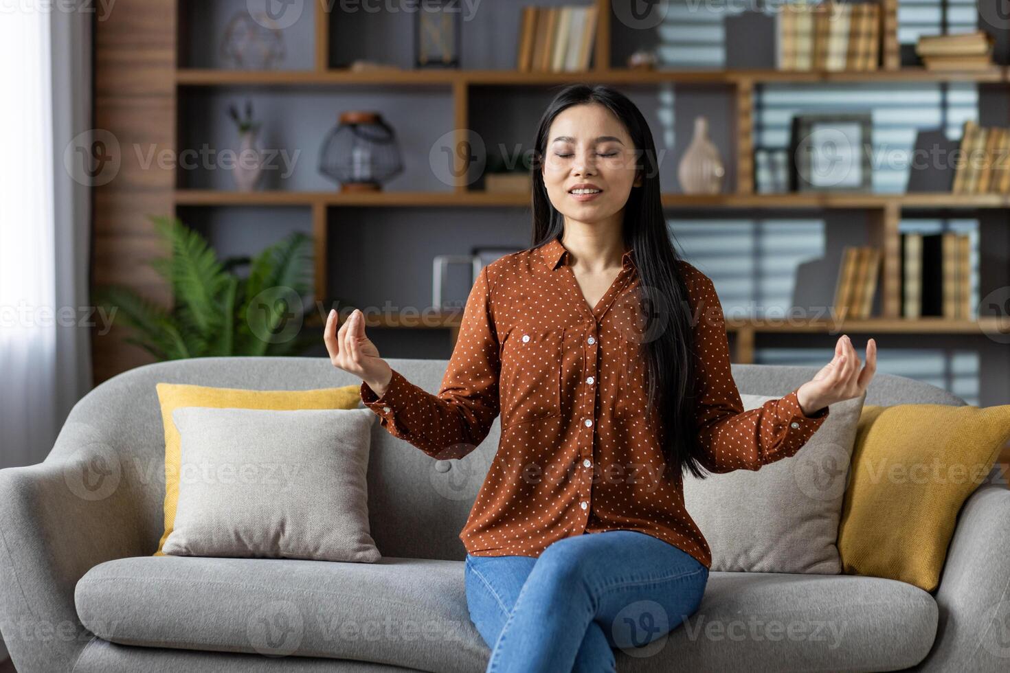 Relaxed Asian woman meditating on sofa at home, enjoying peace and tranquility with a serene expression. Engaged in mindfulness and self-care, embodying calm and relaxation. photo