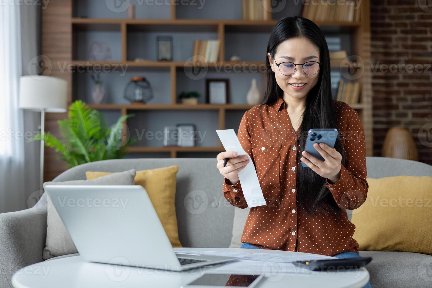 Asian woman managing personal finances from home with smartphone and laptop. Relaxed atmosphere conveys productivity, focus, and efficiency. Ideal for concepts of remote work and technology. photo
