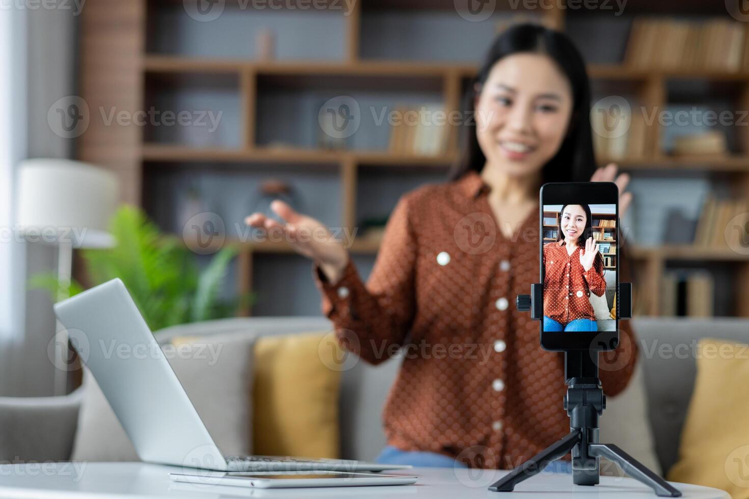 Asian woman using smartphone on tripod at home, recording vlog. Smiling, engaging, laptop visible, sharing ideas. Perfect for illustrating digital content creation, at-home productivity. photo