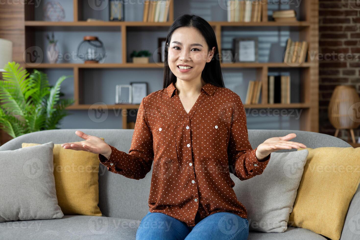 Cheerful asian woman sitting on cozy sofa sharing friendly conversation at home. Relaxed atmosphere with plants, books all around. Positive expression, open gesture, casual clothing. photo