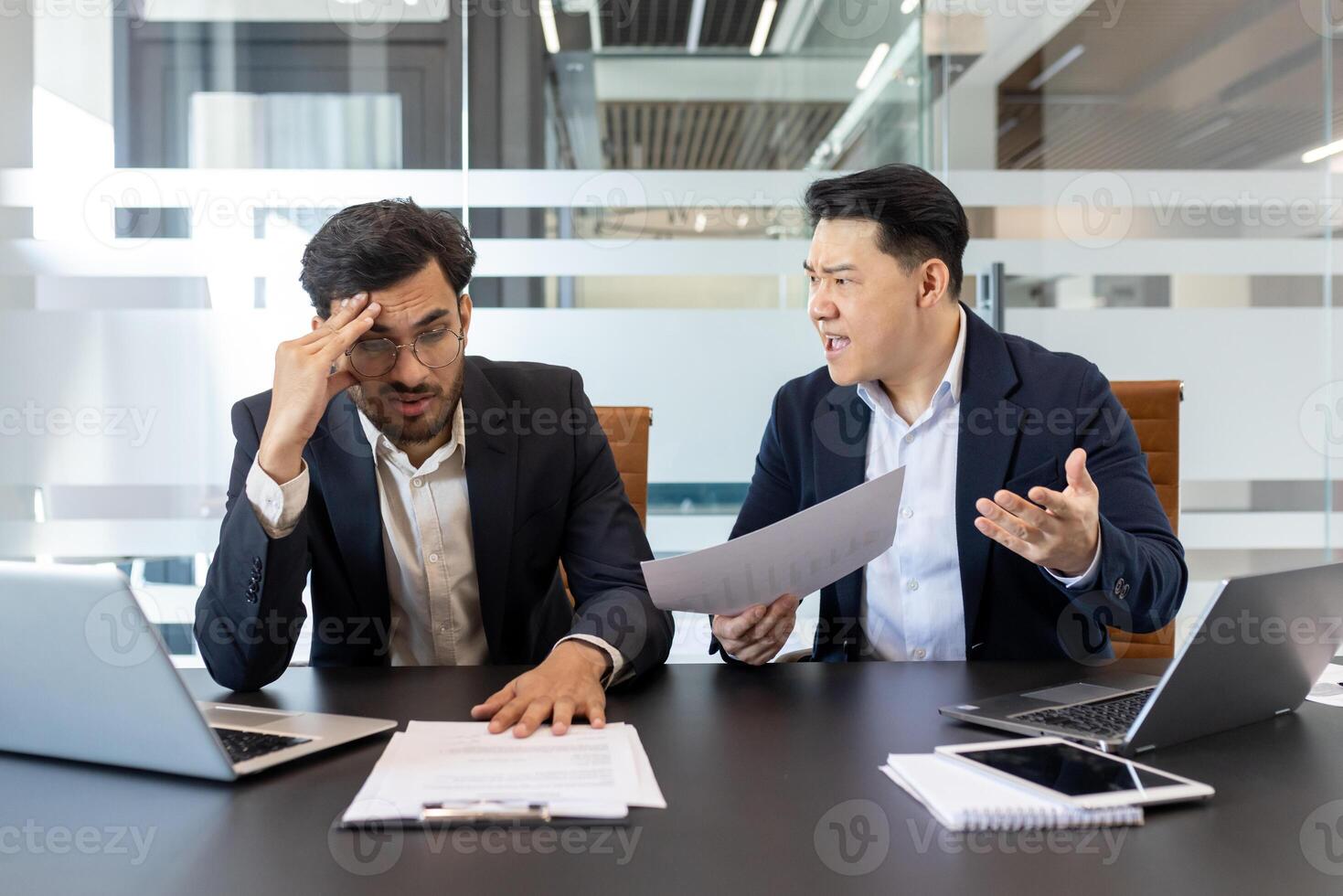 Dissatisfied and frustrated boss yelling at subordinate inside office at workplace. Two men sitting inside office with desk discussing negative report. photo