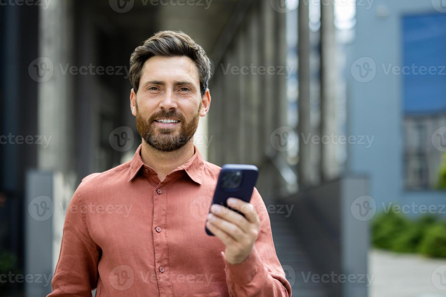 Portrait of successful businessman man with phone in hands. Office worker smiling and looking at camera, using app on smartphone, walking in city from outside office building. photo