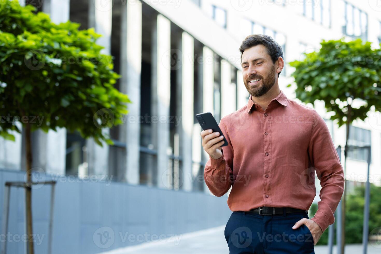 Successful man walking through the city outside an office building with a phone in his hands. Businessman using an app on his smartphone, typing messages, and browsing social media, smiling. photo
