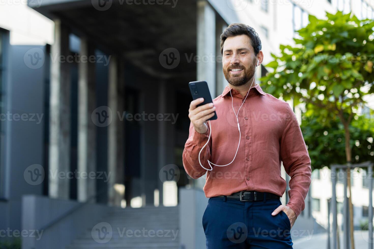 Businessman using wired headphones talking to colleagues and partners while walking around the city. Man smiling contentedly outside office building. photo