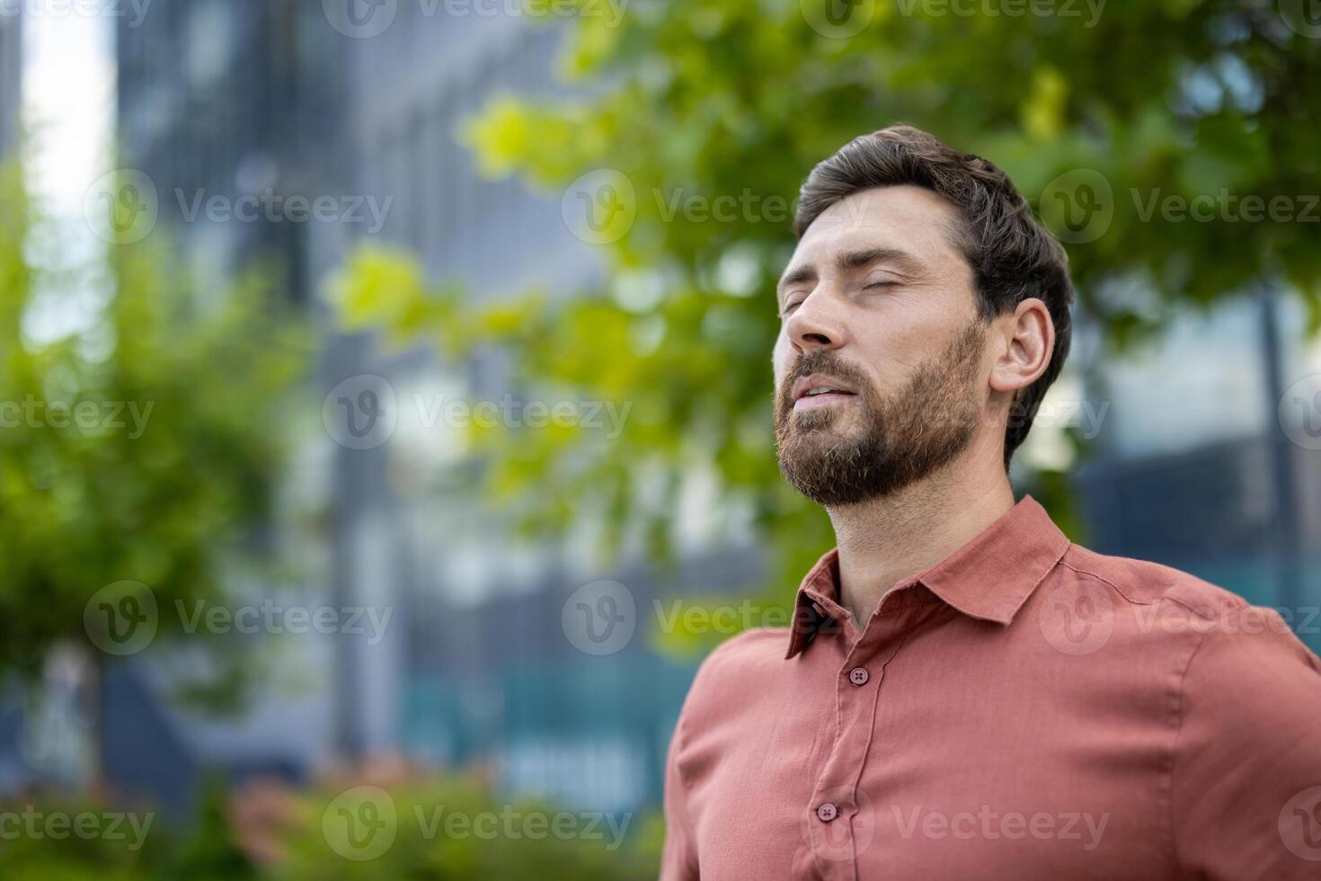 Mature male professional standing outside in city with eyes closed, experiencing calm and relaxation. Businessman taking break, capturing serene and positive emotions in urban setting a photo