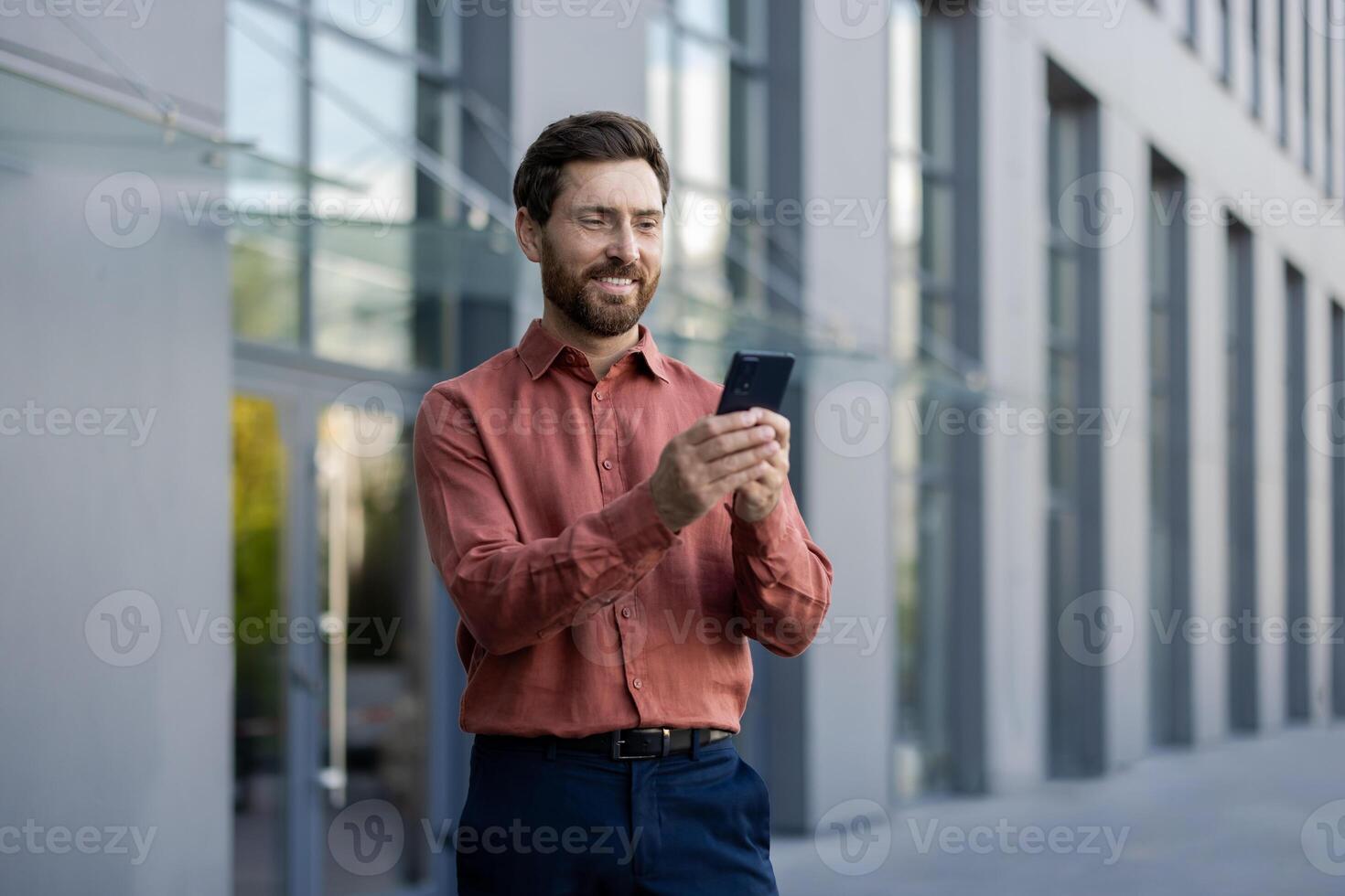 Mature male professional standing outside in city setting, holding phone. Man wearing casual attire, engaging with smartphone. Depicts business, communication, technology, and lifestyle concepts. photo