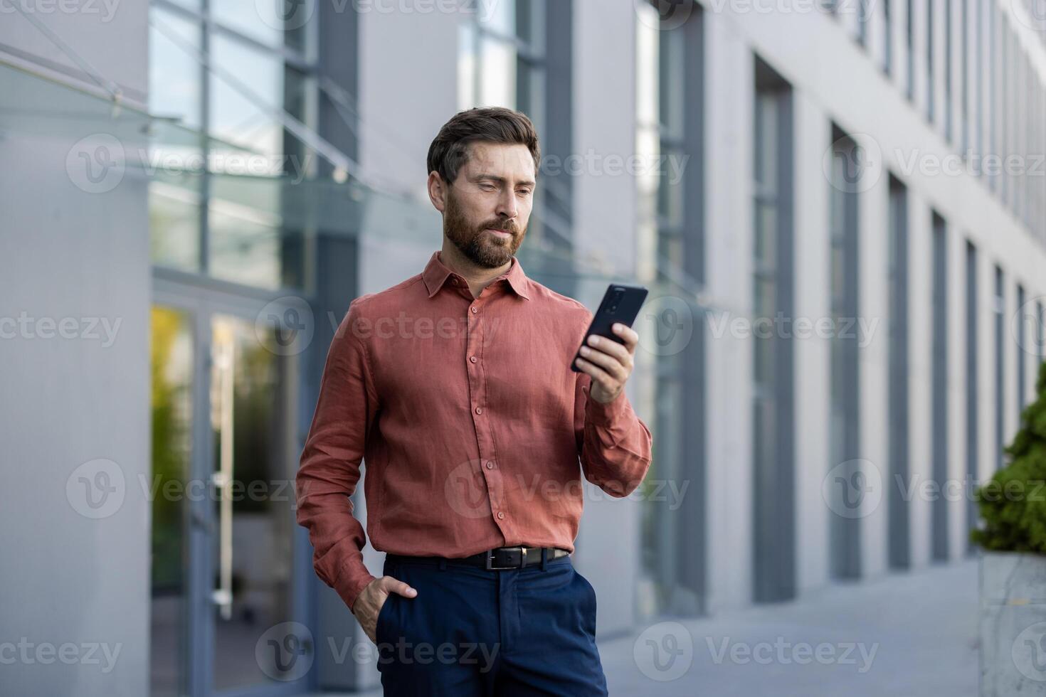 A man in a red shirt is looking at his cell phone. He is standing in front of a building photo