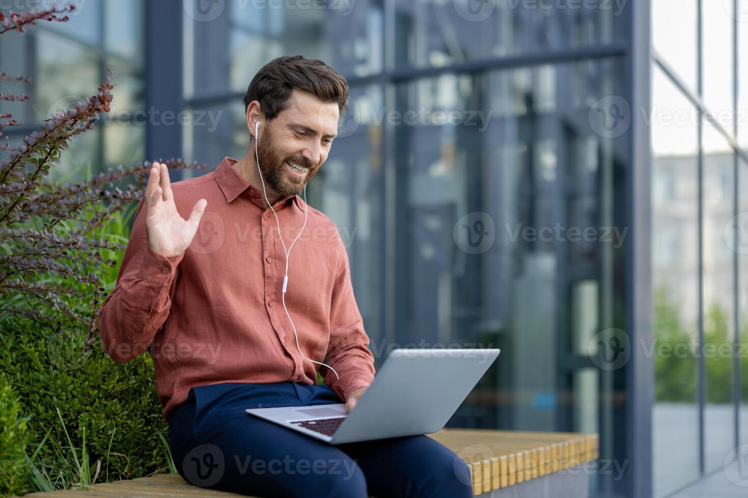 Mature man with earphones using laptop outdoors, waving during call. Engaged in remote work or virtual meeting, showing friendly expression. Urban setting with modern glass building. photo