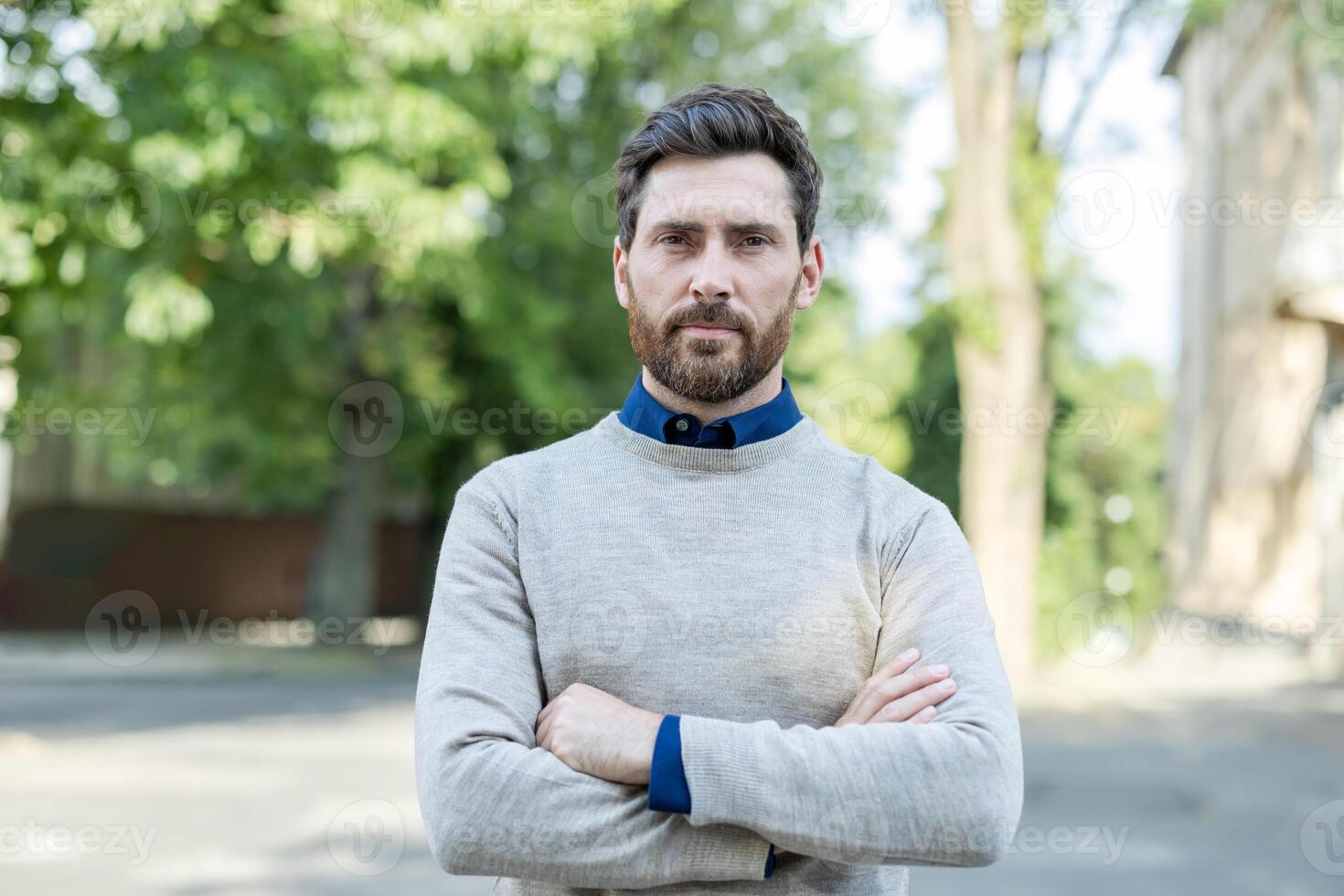 Portrait of confident mature man with arms crossed standing outdoors. Expresses self-assurance and authority against blurred background of trees and buildings. Casual and poised demeanor. photo