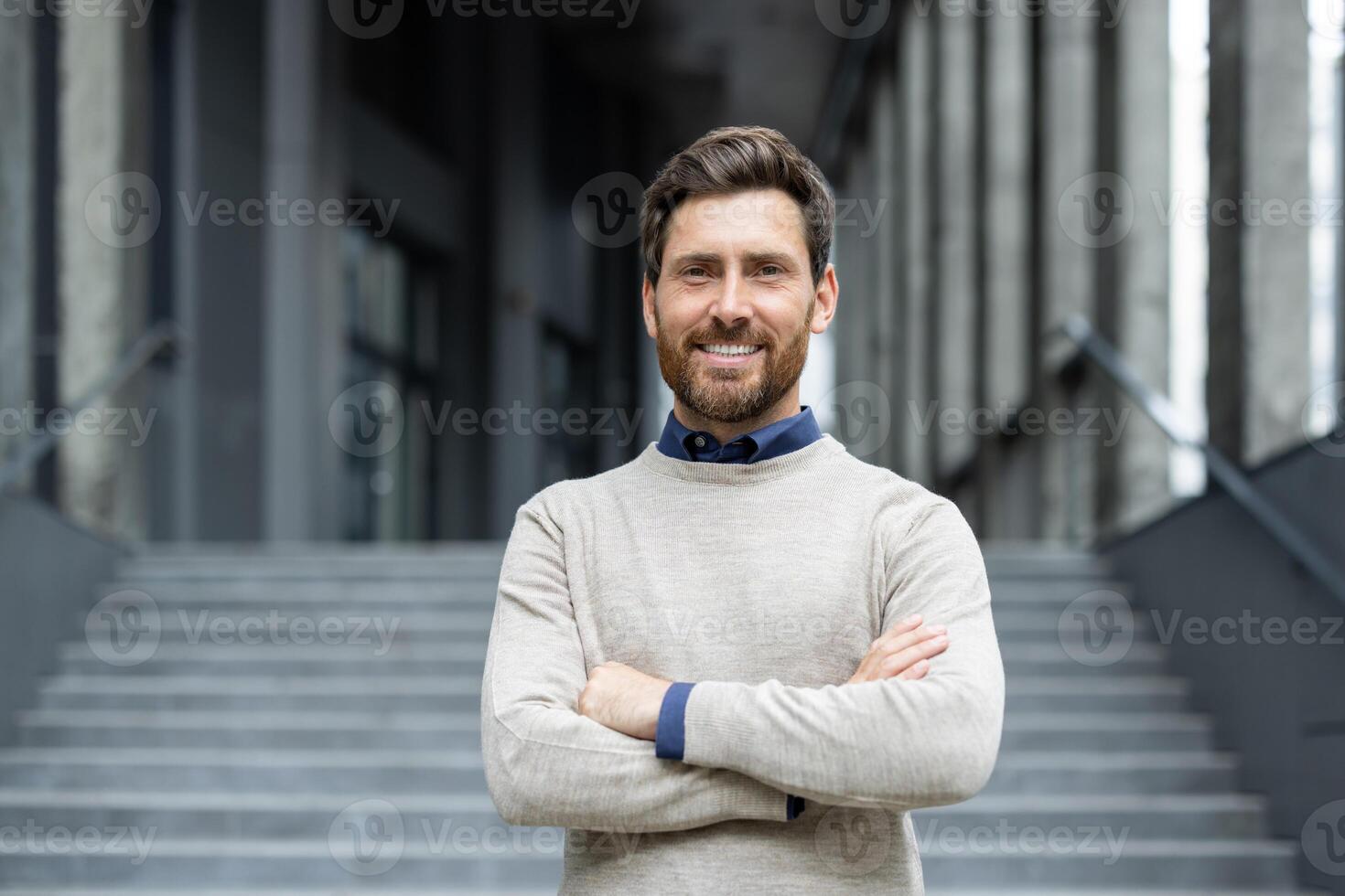 Confident businessman standing with arms crossed outside modern office building. Wearing casual sweater, showcasing professional demeanor success. Smiling, relaxed ambiance conveys approachability photo