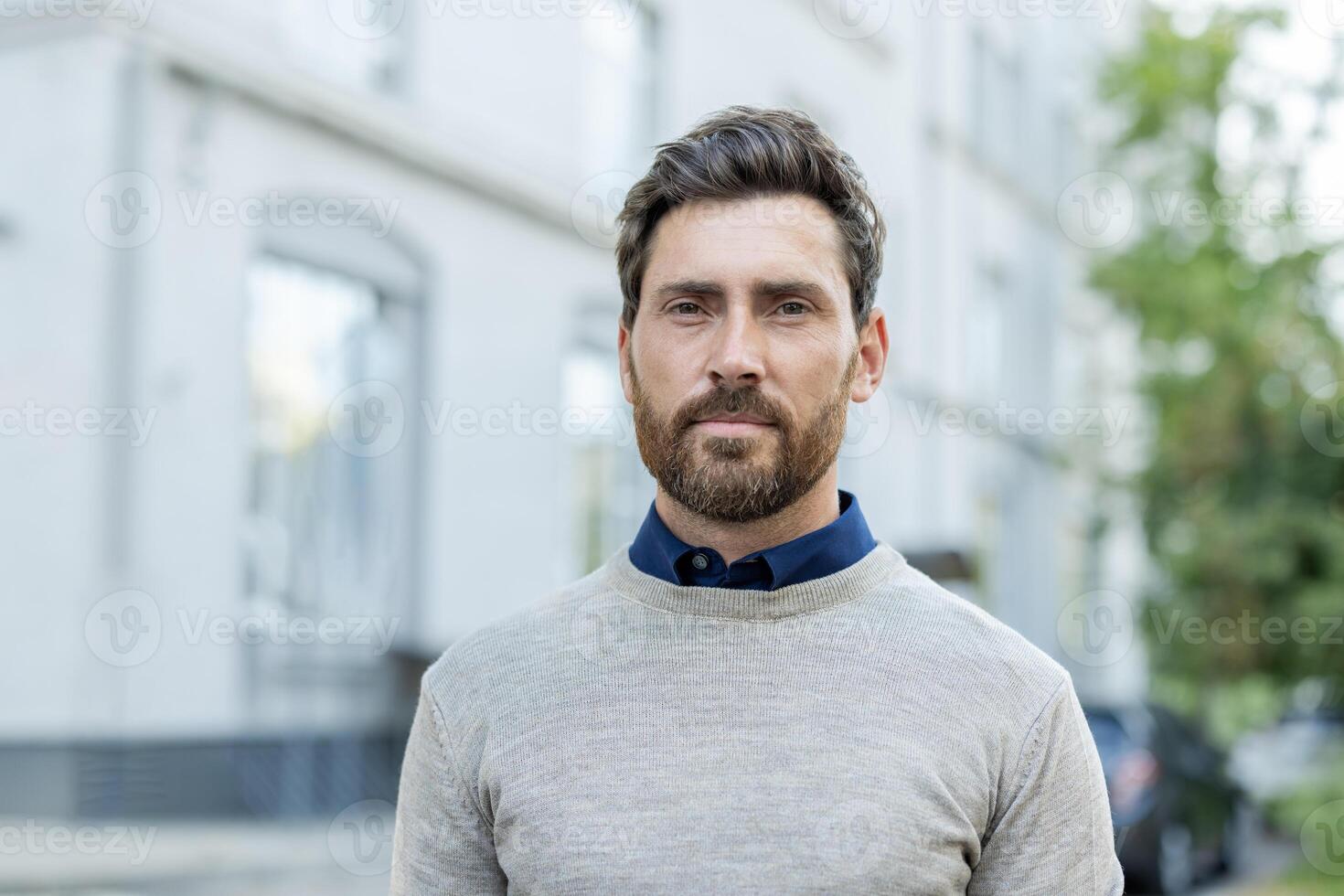A man with a beard and a blue shirt is standing in front of a building. He is wearing a gray sweater photo