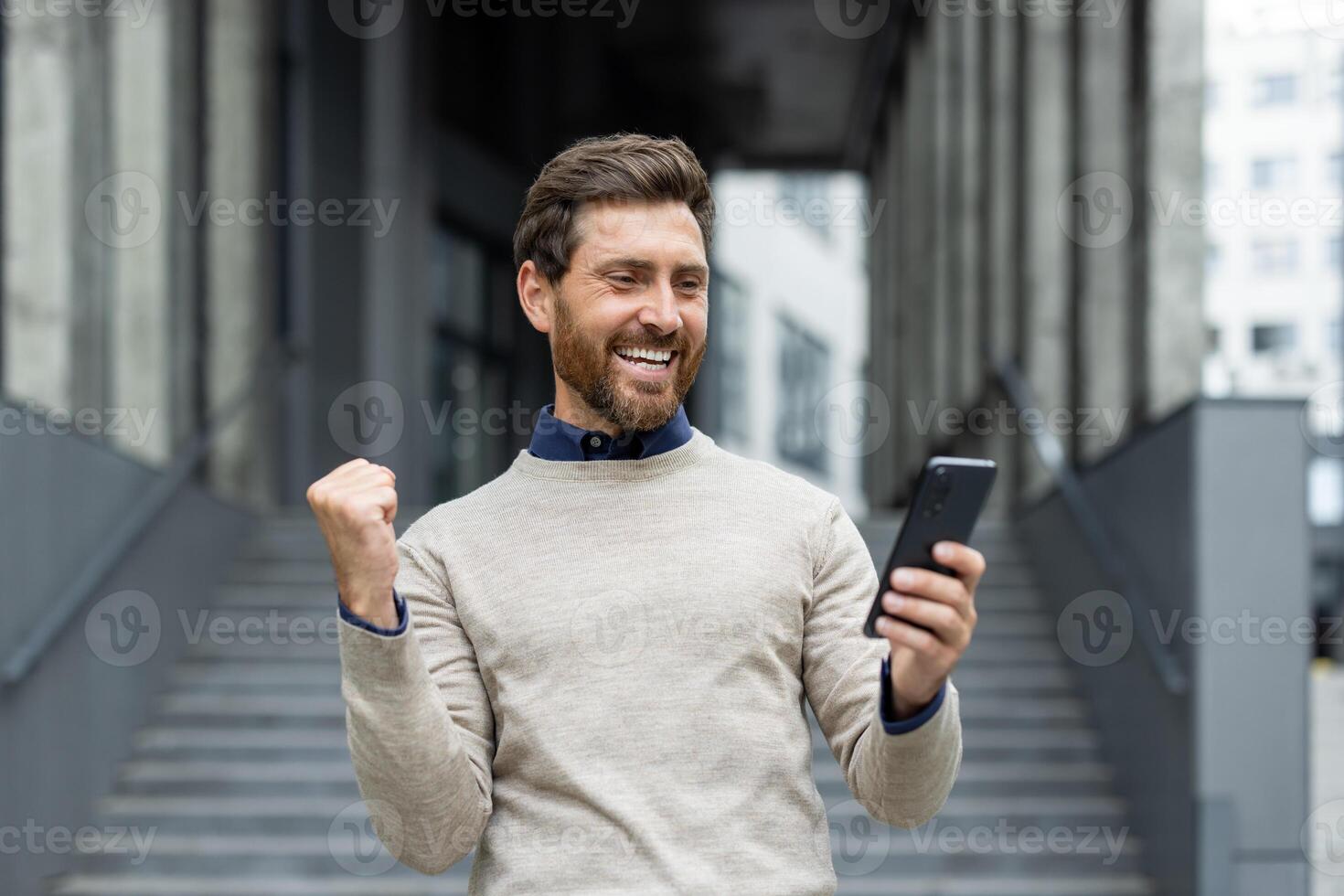 Smiling man with beard holding phone celebrating positive news or achievement. Expressing joy and success in urban office setting, displaying emotions of excitement and happiness. photo