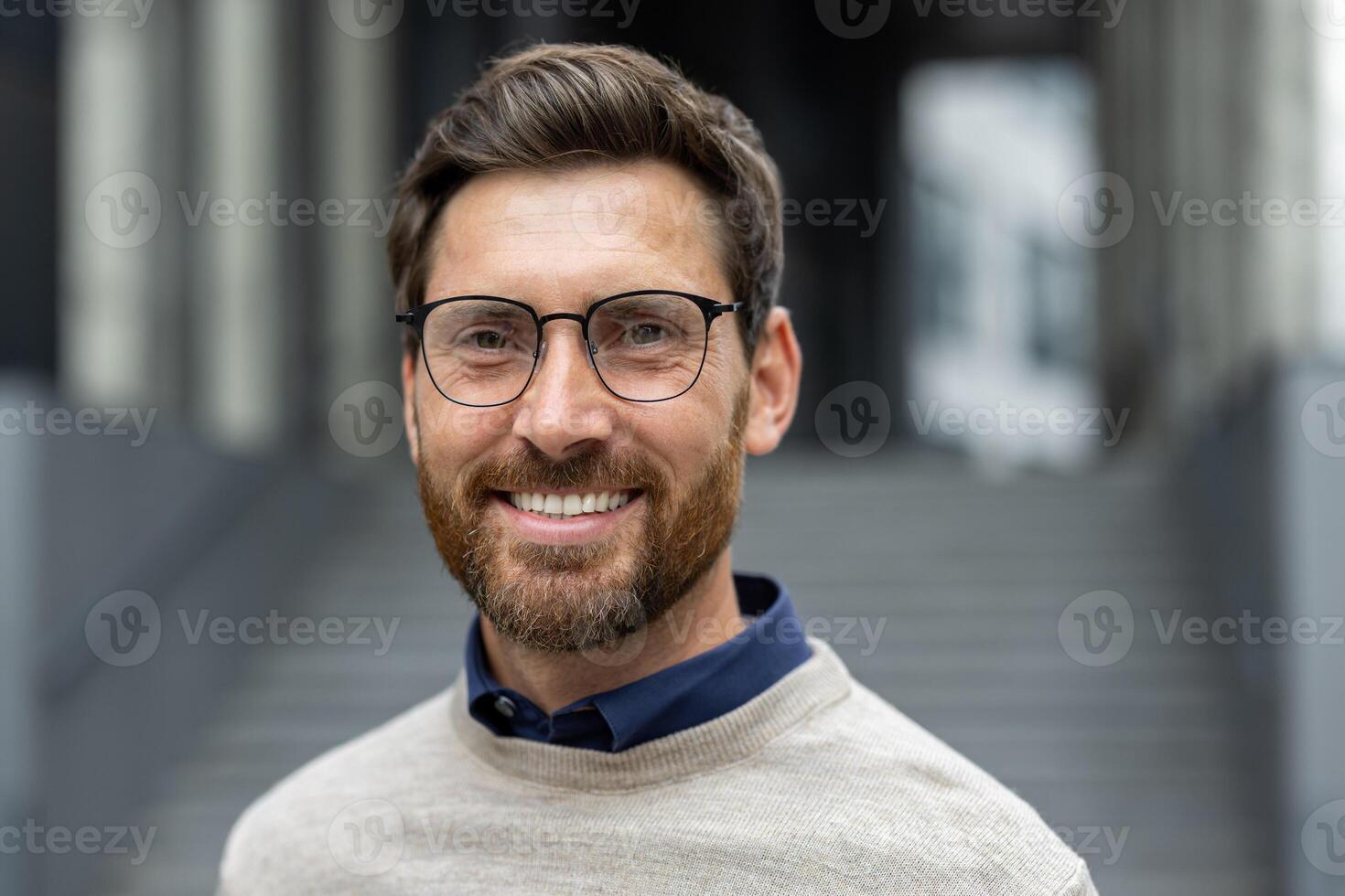 Portrait of confident man with glasses smiling warmly, conveying approachability and professionalism. Wearing stylish sweater, he stands in urban environment, suggesting success and positivity in business or teaching professions. photo