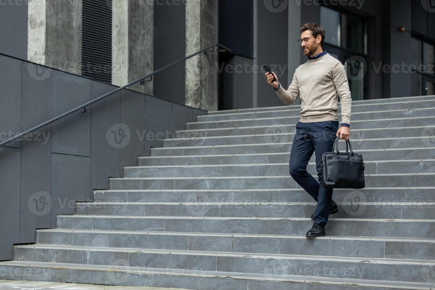 Businessman walking down grey concrete steps using smartphone, dressed in smart casual outfit, holding black leather briefcase. Image represents professional urban lifestyle, confidence, communication, mobility in business environment. photo