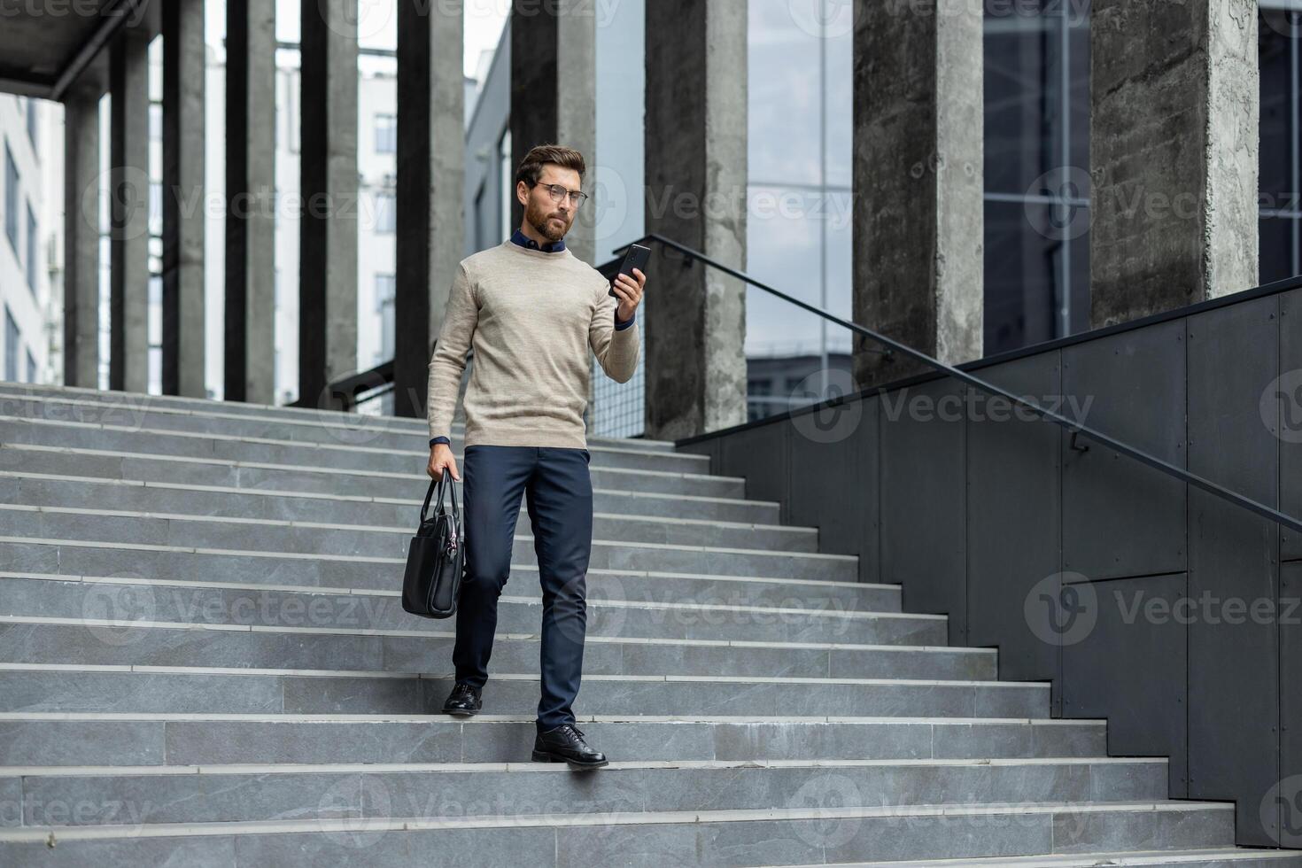 Business professional in modern outfit walking down city stairs using phone for important communication. Man looks focused, balancing work and mobile engagement, holding briefcase photo