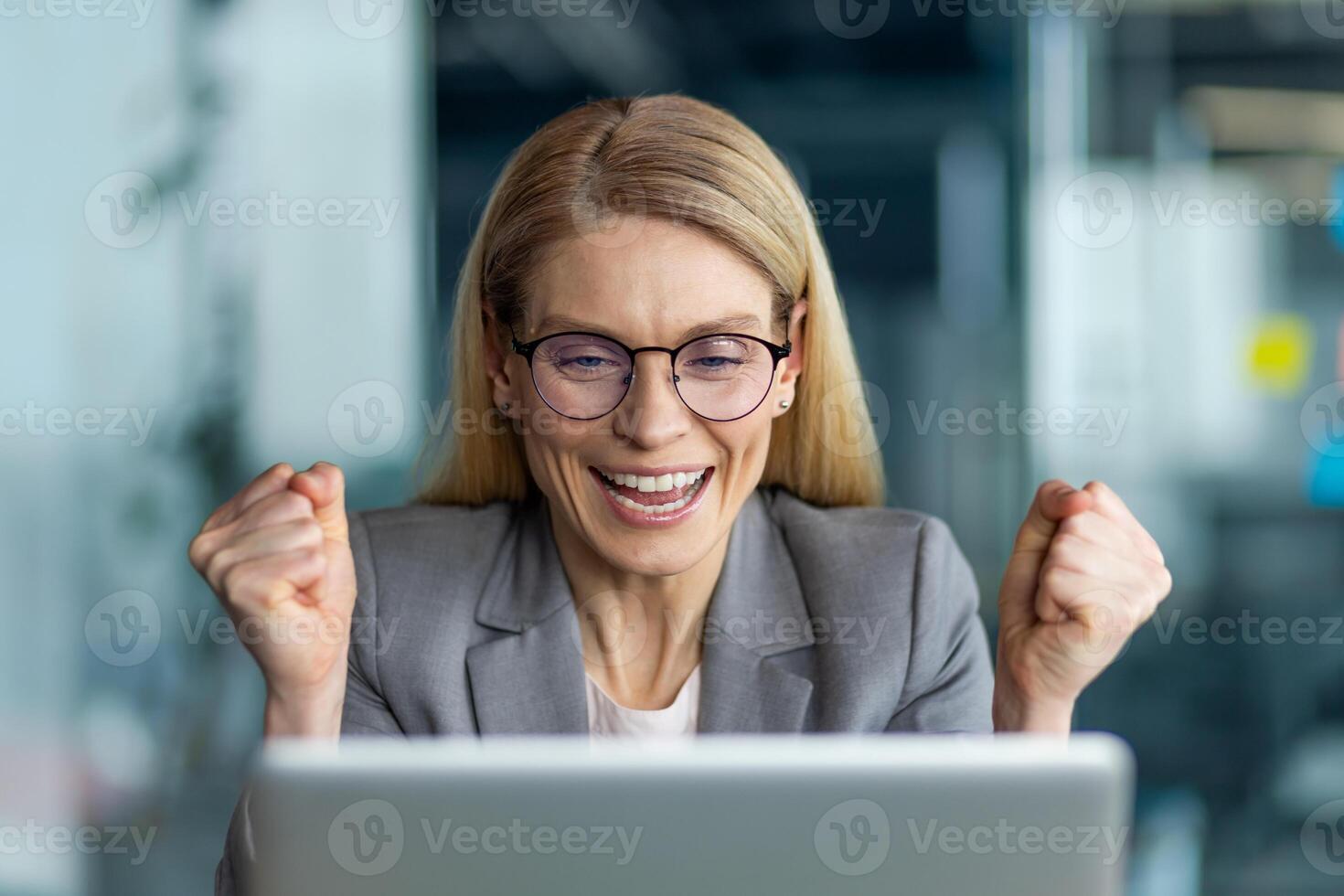Businesswoman expressing joy and excitement while working on laptop in office. Professional in glasses and blazer displaying triumphant gesture, highlighting success and achievement in business photo