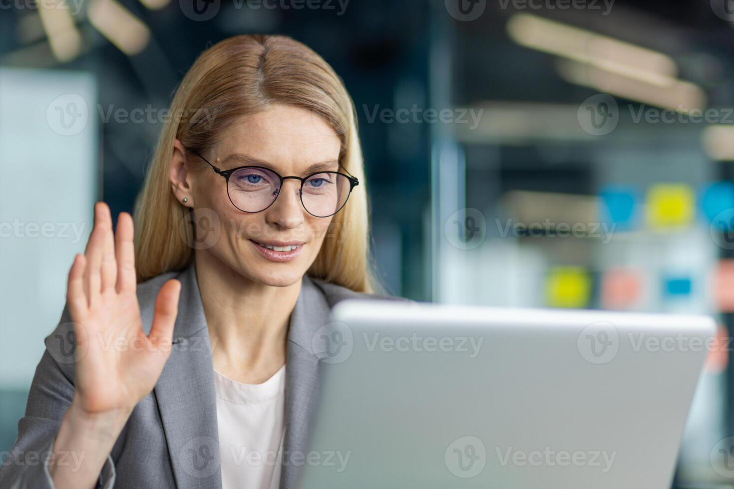 Professional woman in office using laptop for call. She waves hand at screen, engaged in virtual communication. Glasses and suit reflect work environment. Technology enhances teleconference. photo