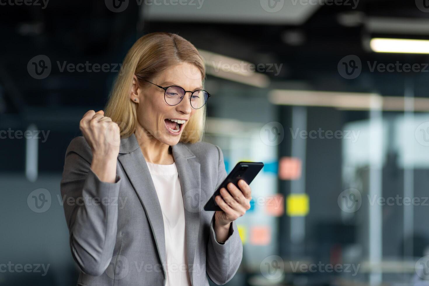 Businesswoman in office environment celebrates success with phone in hand, expressing joy and excitement. Demonstrates achievement, happiness, and motivation in professional setting. photo