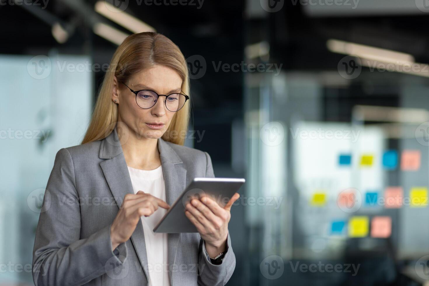 Confident businesswoman in gray suit using digital tablet in bright modern office. She looks focused, embodying professionalism and technology-driven work environment. photo