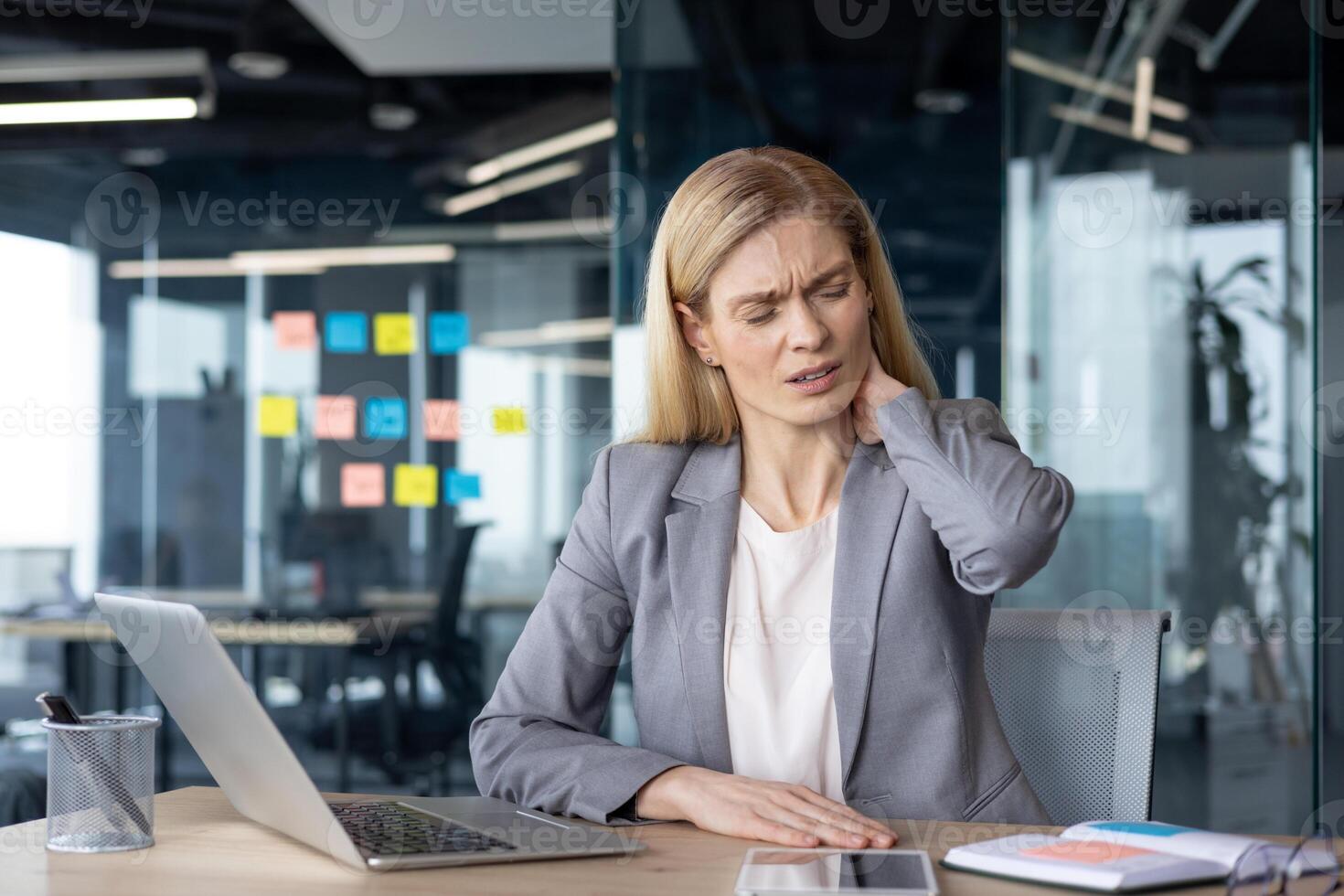 Mature businesswoman working on laptop at an office , expressing noticeable neck pain. The scene highlights workplace discomfort and ergonomic challenges faced by professionals in busy environment. photo
