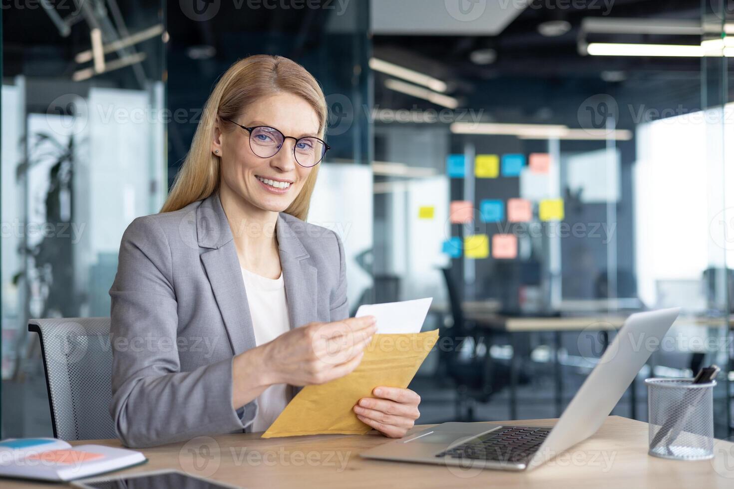 Mature woman businesswoman holding an envelope and smiling while working in a contemporary office. The workspace features a laptop and organizational tools, creating a productive work setting. photo