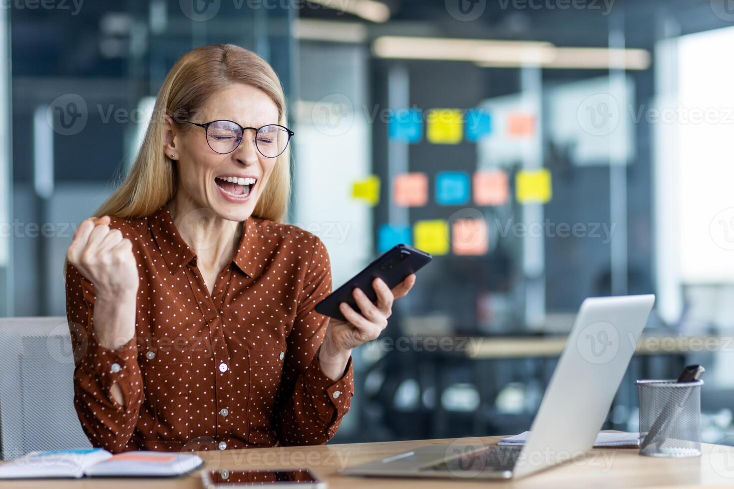 Joyful businesswoman sits in office using phone to celebrate professional success. Enthusiastic expression shows positivity. Laptop and sticky notes signify productive work environment photo