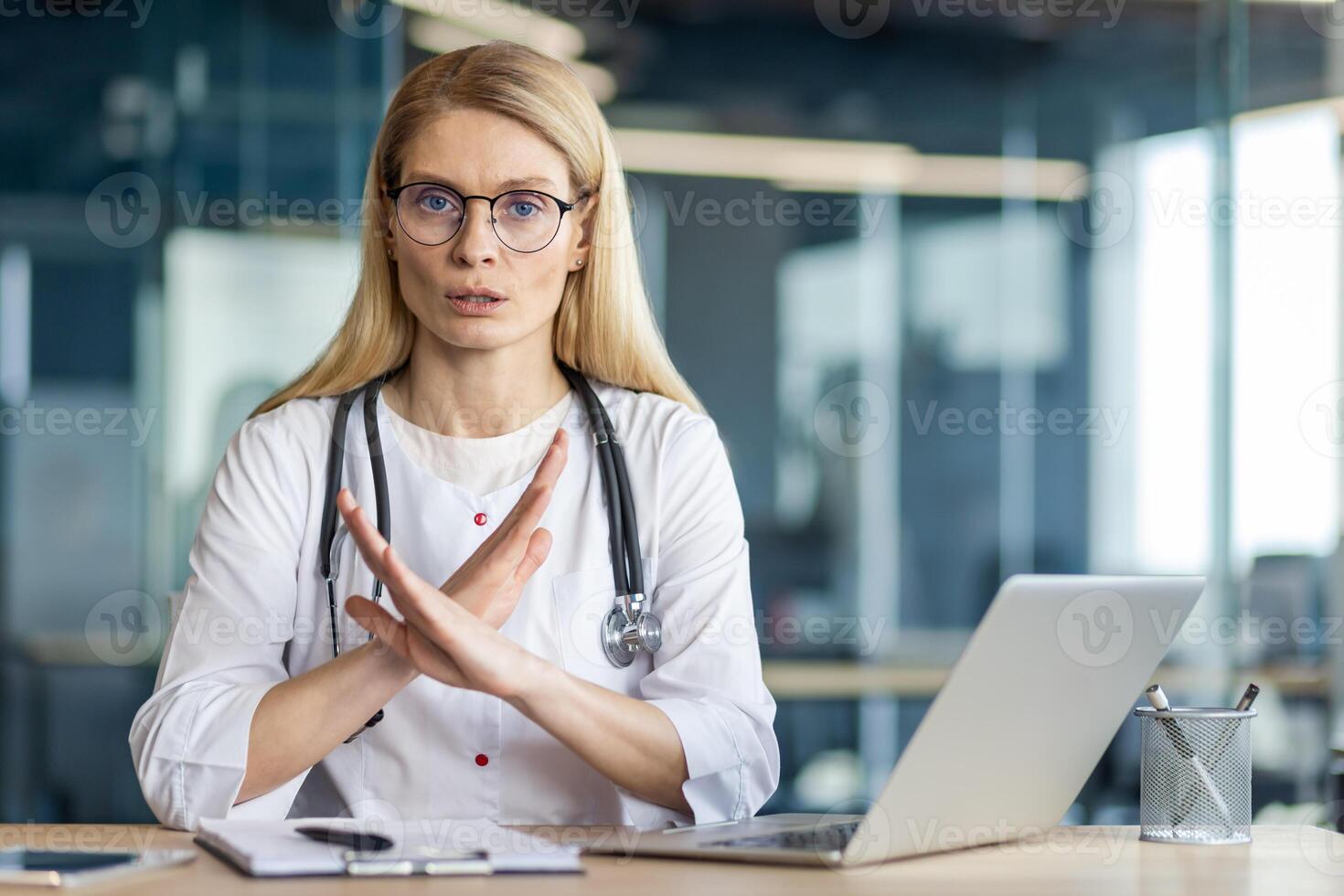 Female doctor in white coat with stethoscope gestures no at laptop desk. She wears glasses, communicates confidently in modern office, expressing authority and professionalism. photo