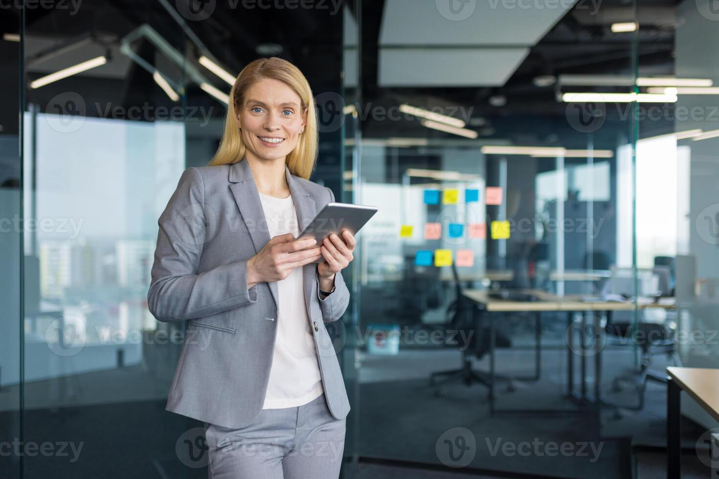 Portrait of a successful mature businesswoman with a tablet computer in her hands. Office worker in a business suit smiling and looking at the camera, standing near the window inside the office. photo