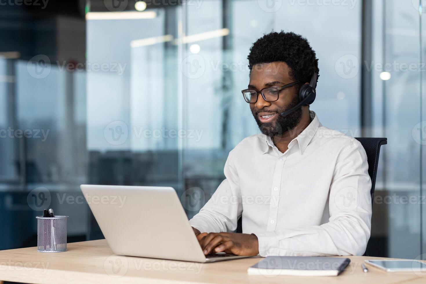 African American man seated at workplace using laptop and headset for communication. Displays focus, technology, remote work efficiency. Ideal for modern business concepts and professional settings. photo