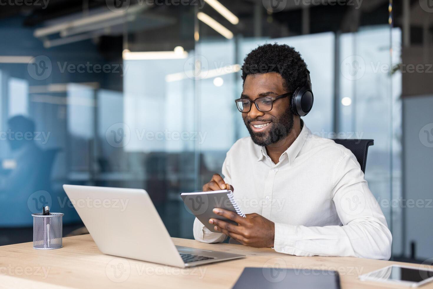 African American businessman engaged in online learning using laptop and notebook. Wearing headphones, he is focused and smiling in modern office environment. photo