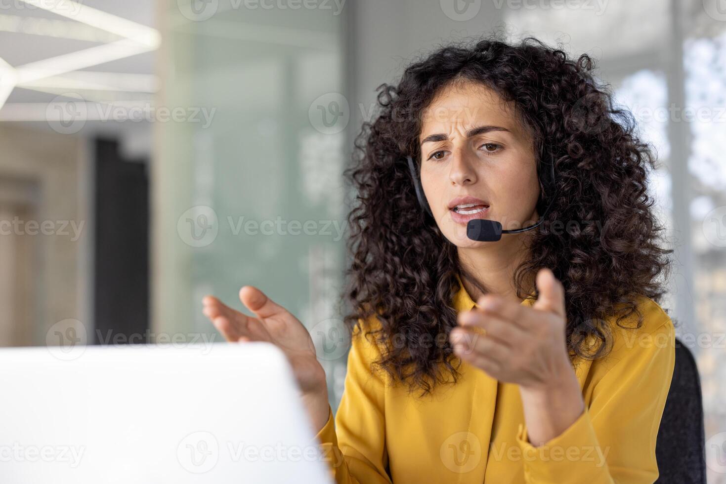 Serious and focused woman with headset talking remotely, close-up of helpline worker working remotely, using laptop for call, at workplace inside office. photo