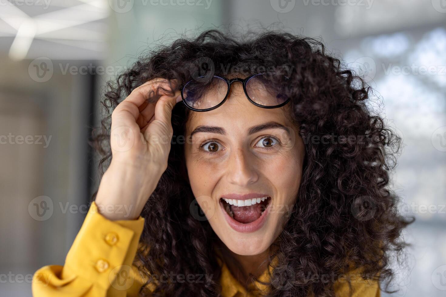 Hispanic businesswoman showcasing surprise and excitement in office, lifting glasses with cheerful expression. Captures moment of joy and enthusiasm in professional environment. photo